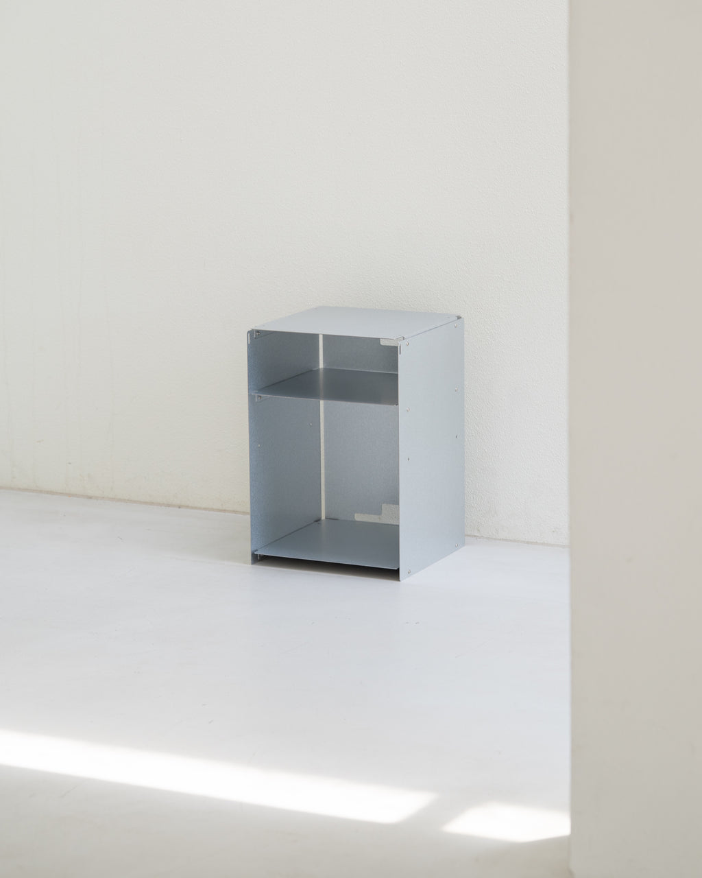 A minimalist, open-top metal storage cabinet in a light gray finish, set against a plain white wall and floor with soft natural light casting a diagonal shadow.
