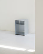 A minimalist, open-top metal storage cabinet in a light gray finish, set against a plain white wall and floor with soft natural light casting a diagonal shadow.