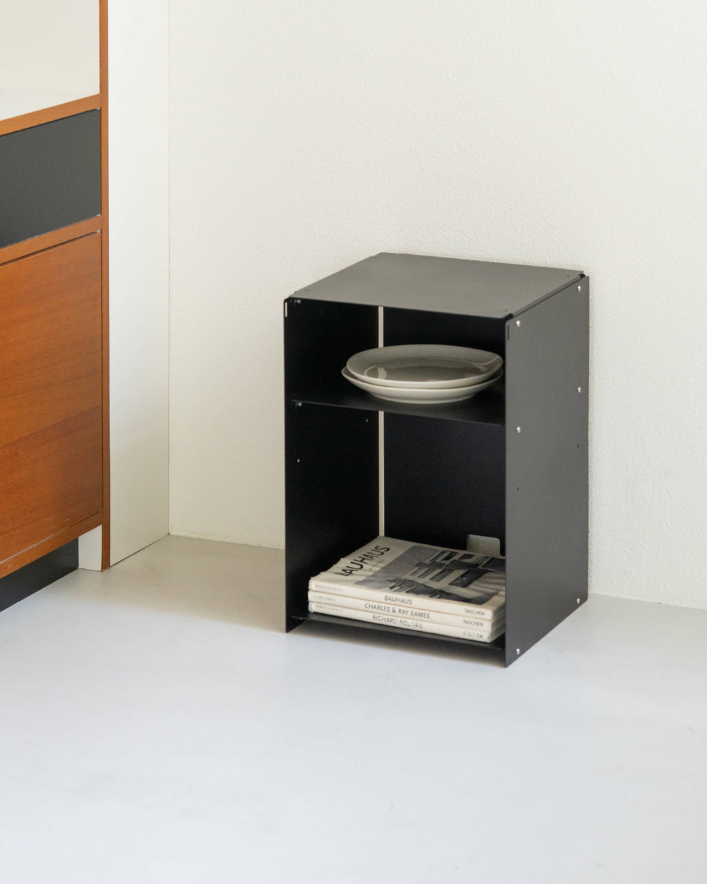 A small black metal storage cabinet with two open shelves, shown in a minimalist room corner. The top shelf holds a stack of gray plates, and the bottom shelf holds a stack of magazines.