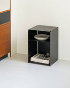 A small black metal storage cabinet with two open shelves, shown in a minimalist room corner. The top shelf holds a stack of gray plates, and the bottom shelf holds a stack of magazines.