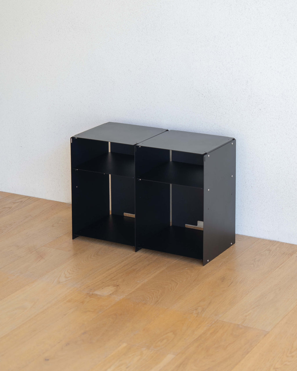 Two small black modular storage boxes with open shelves, placed on a light wood floor against a white wall.