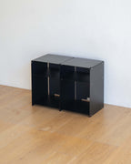 Two small black modular storage boxes with open shelves, placed on a light wood floor against a white wall.