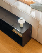 A modern black modular side table with a small white table lamp on top, positioned next to a light beige sofa on a wooden floor.
