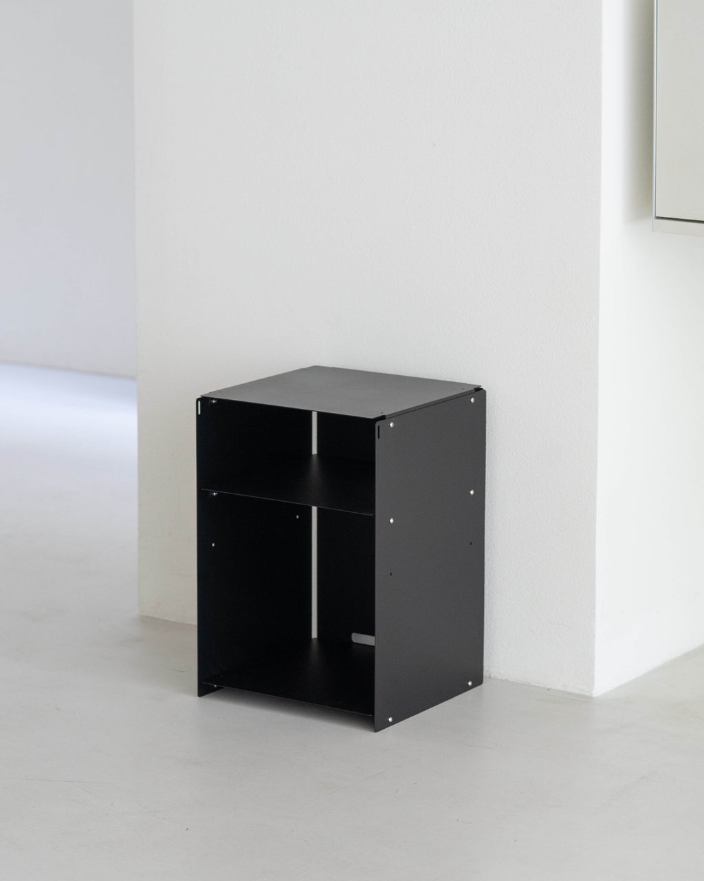 A minimalist black metal two-tier storage cabinet with open shelves, positioned in a clean, modern white room corner.