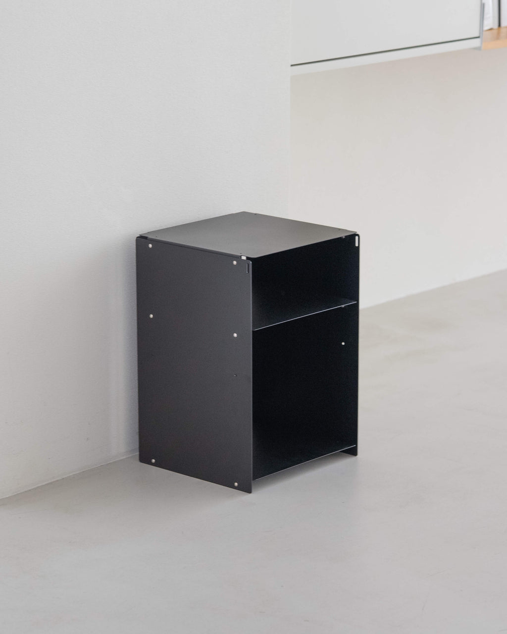 A minimalist black metal side table with an open top and one open shelf, positioned in a clean, modern room corner with light gray concrete floors and white walls.