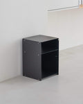 A minimalist black metal side table with an open top and one open shelf, positioned in a clean, modern room corner with light gray concrete floors and white walls.