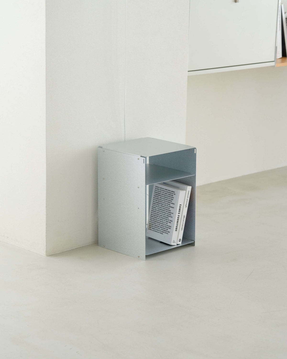 A minimalist light gray open storage box or bookshelf holding a few books, placed in the corner of a bright, modern room with white walls and a light concrete floor.