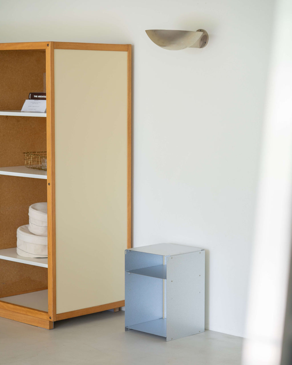 A minimalist interior scene showing a tall wooden cabinet with a cream-colored door and open shelving, alongside a small light blue side table against a white wall with a sculptural wall lamp.