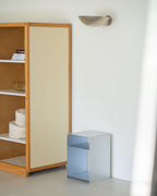 A minimalist interior scene showing a tall wooden cabinet with a cream-colored door and open shelving, alongside a small light blue side table against a white wall with a sculptural wall lamp.