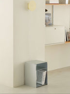 A minimalist interior scene with a small open metal storage box on the floor containing books, a wall-mounted shelf with decor, and a round wall light above.