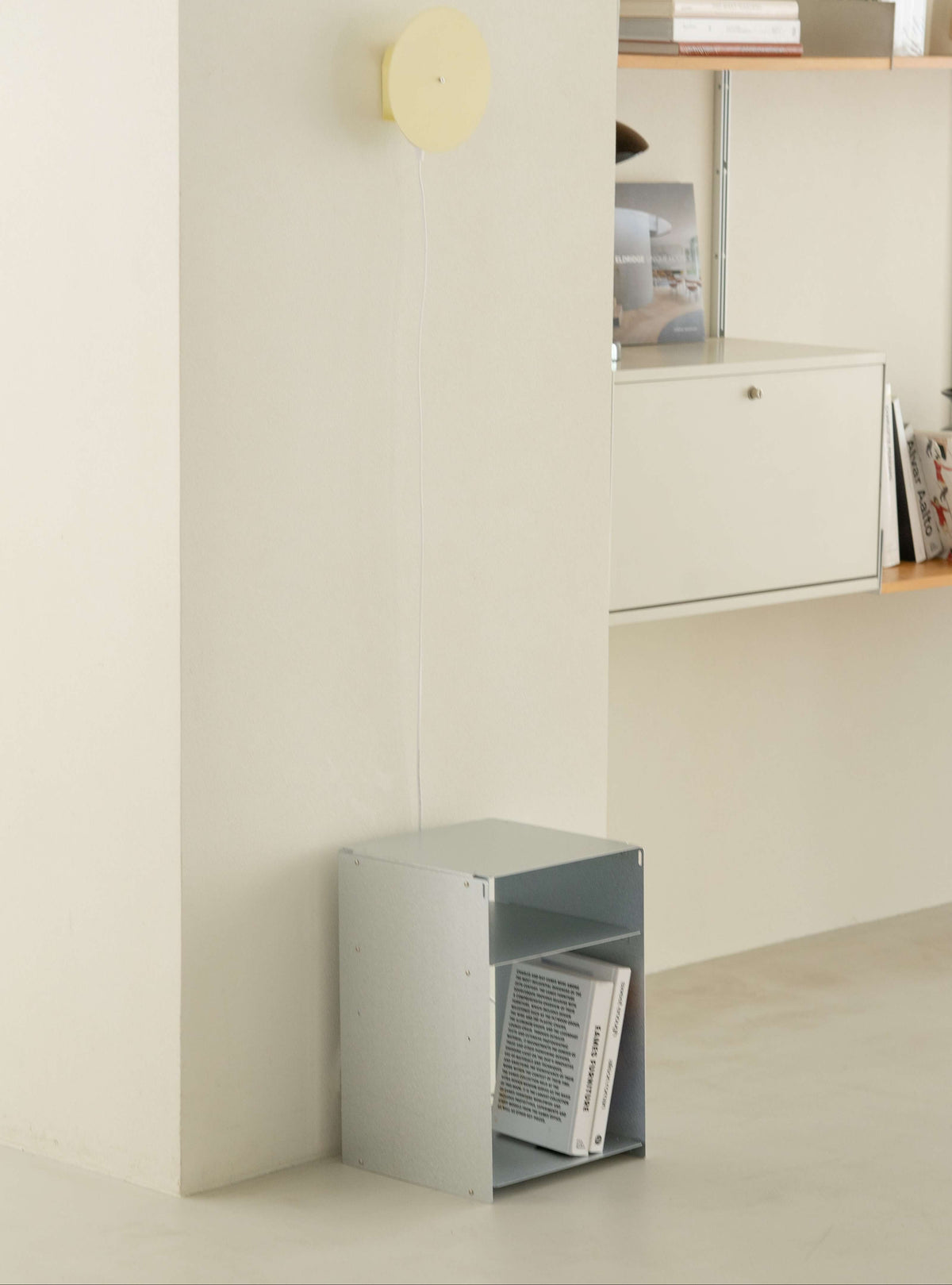 A minimalist interior scene with a small open metal storage box on the floor containing books, a wall-mounted shelf with decor, and a round wall light above.