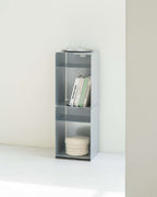 A tall, narrow gray metal shelving unit with three open compartments, styled with books and a round tray on top, set against a minimalist off-white wall and floor.