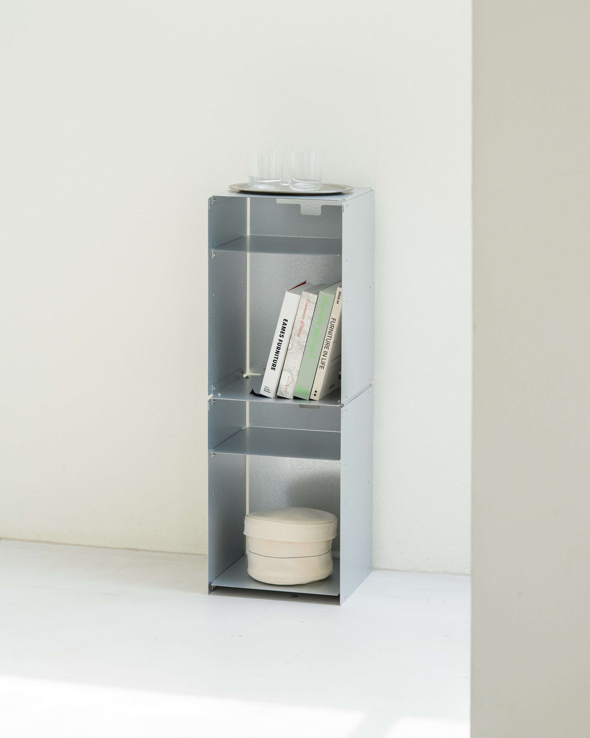 A tall, narrow gray metal shelving unit with three open compartments, styled with books and a round tray on top, set against a minimalist off-white wall and floor.