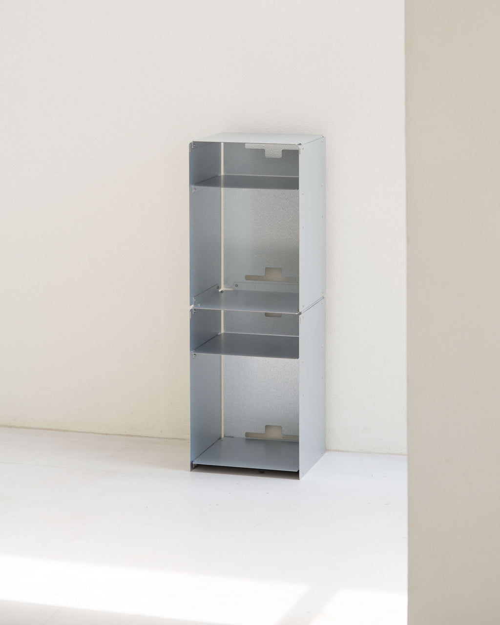 A tall, minimalist three-tier metal shelving unit in a light gray finish, set against a clean, off-white minimalist wall and floor.