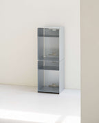A tall, minimalist three-tier metal shelving unit in a light gray finish, set against a clean, off-white minimalist wall and floor.