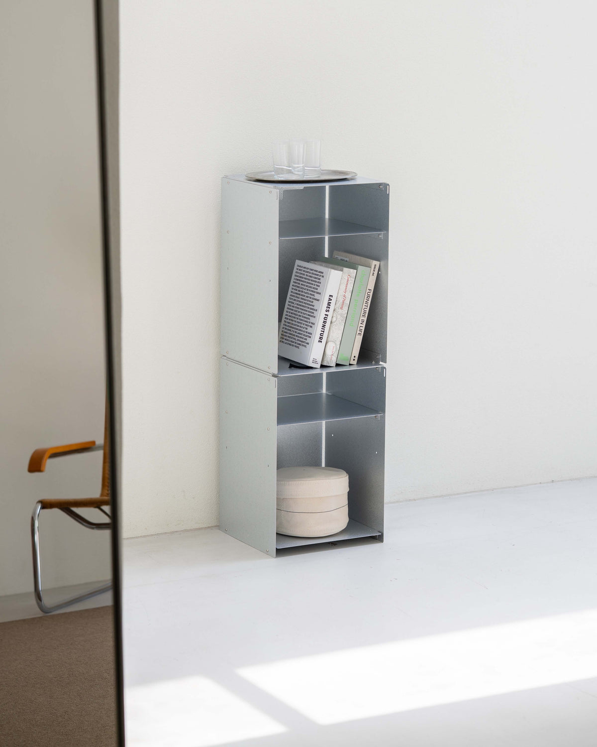A tall, narrow open shelving unit with three tiers of open cubbies, shown in a minimalist room corner. The unit is light gray and contains a few books and a round container on top.