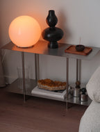 A modern two-tier metal side table styled with a glowing orange globe lamp, a black geometric vase, a small incense holder, and other decor, set in a cozy room corner.