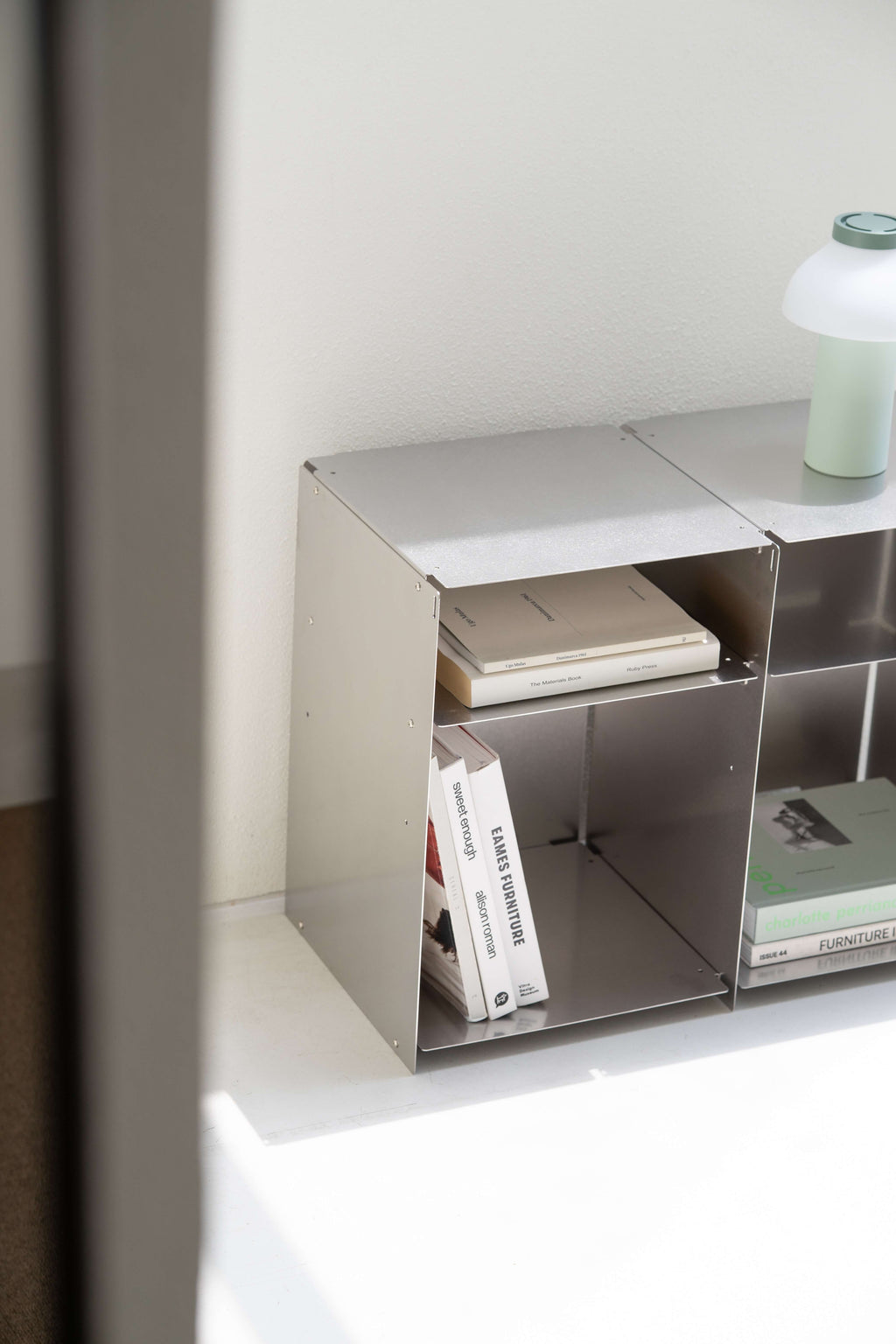 A minimalist silver metal modular shelf organizer holding books and a small green lamp, set against a clean white wall.