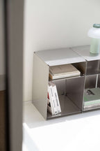 A minimalist silver metal modular shelf organizer holding books and a small green lamp, set against a clean white wall.