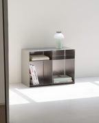 A minimalist metallic gray modular storage shelf with open cubbies, styled with books and a small table lamp, set against a clean off-white wall and floor.