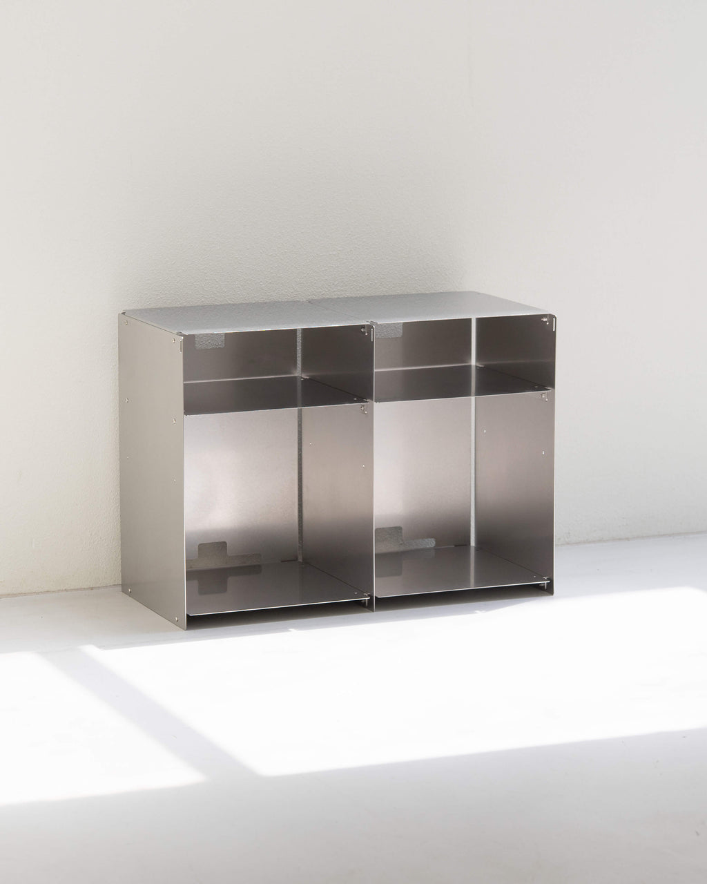 A modern two-compartment stainless steel storage cabinet with open shelves, set against a minimalist white wall and floor with soft natural light casting a shadow.
