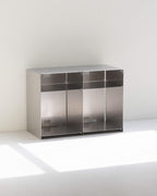 A modern two-compartment stainless steel storage cabinet with open shelves, set against a minimalist white wall and floor with soft natural light casting a shadow.