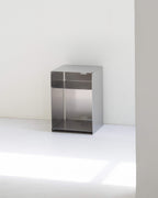 A minimalist stainless steel storage cabinet with a single open top shelf and a lower door, set against a clean white wall and floor.