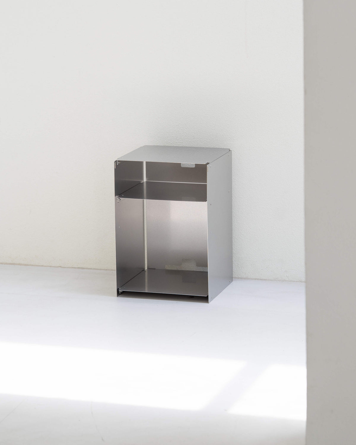 A minimalist stainless steel storage cabinet with a single open top shelf and a lower door, set against a clean white wall and floor.