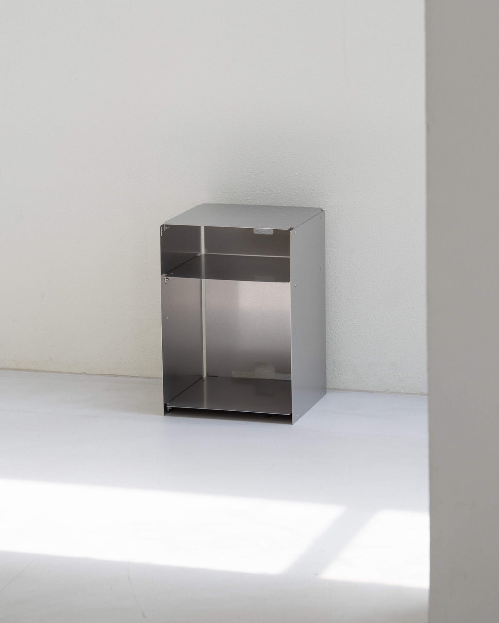 A minimalist, open-top metal storage cabinet with a brushed silver finish, positioned against a plain white wall and floor.