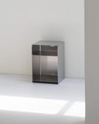 A minimalist, open-top metal storage cabinet with a brushed silver finish, positioned against a plain white wall and floor.