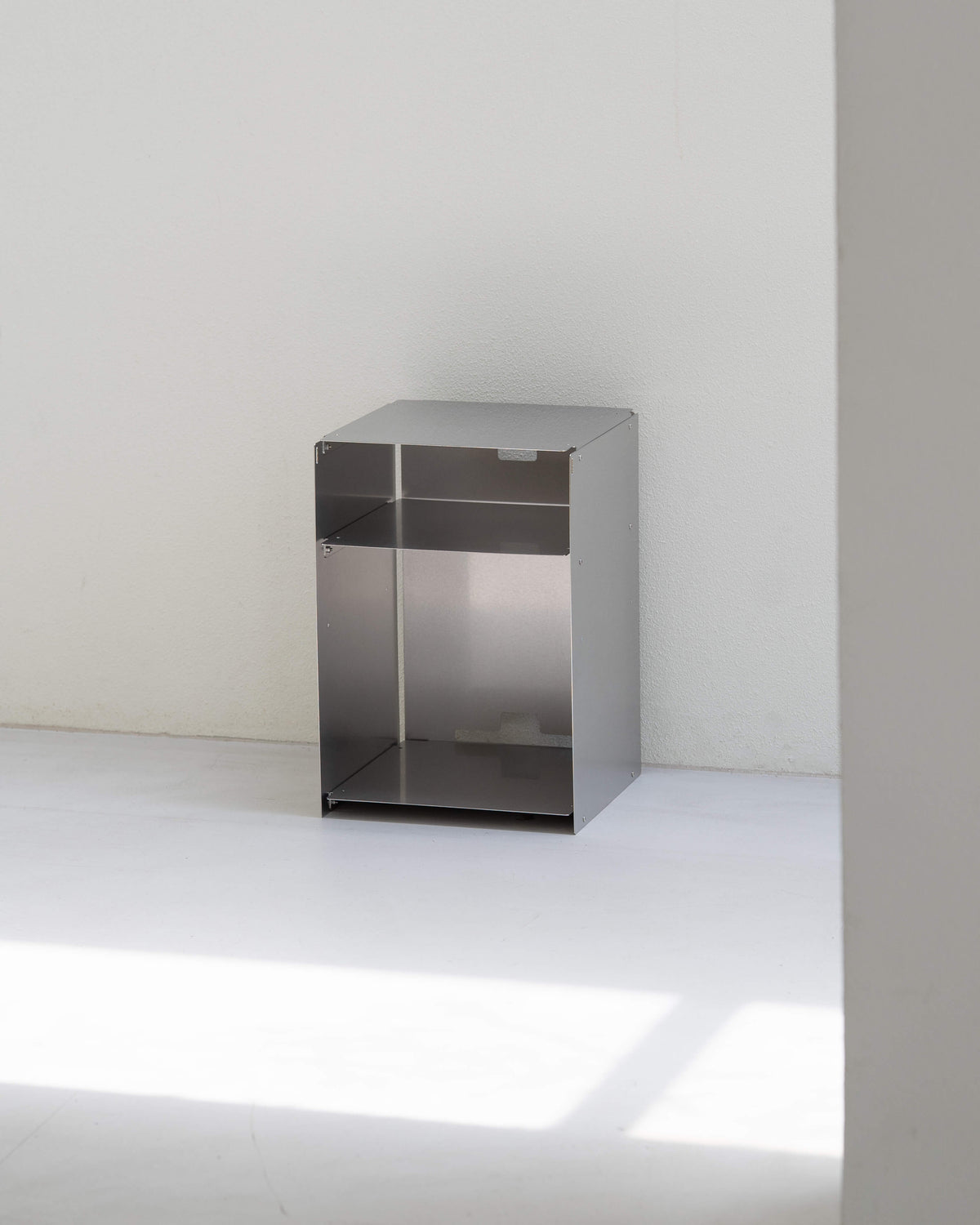 A minimalist, open-top metal storage cabinet with a brushed silver finish, positioned against a plain white wall and floor.