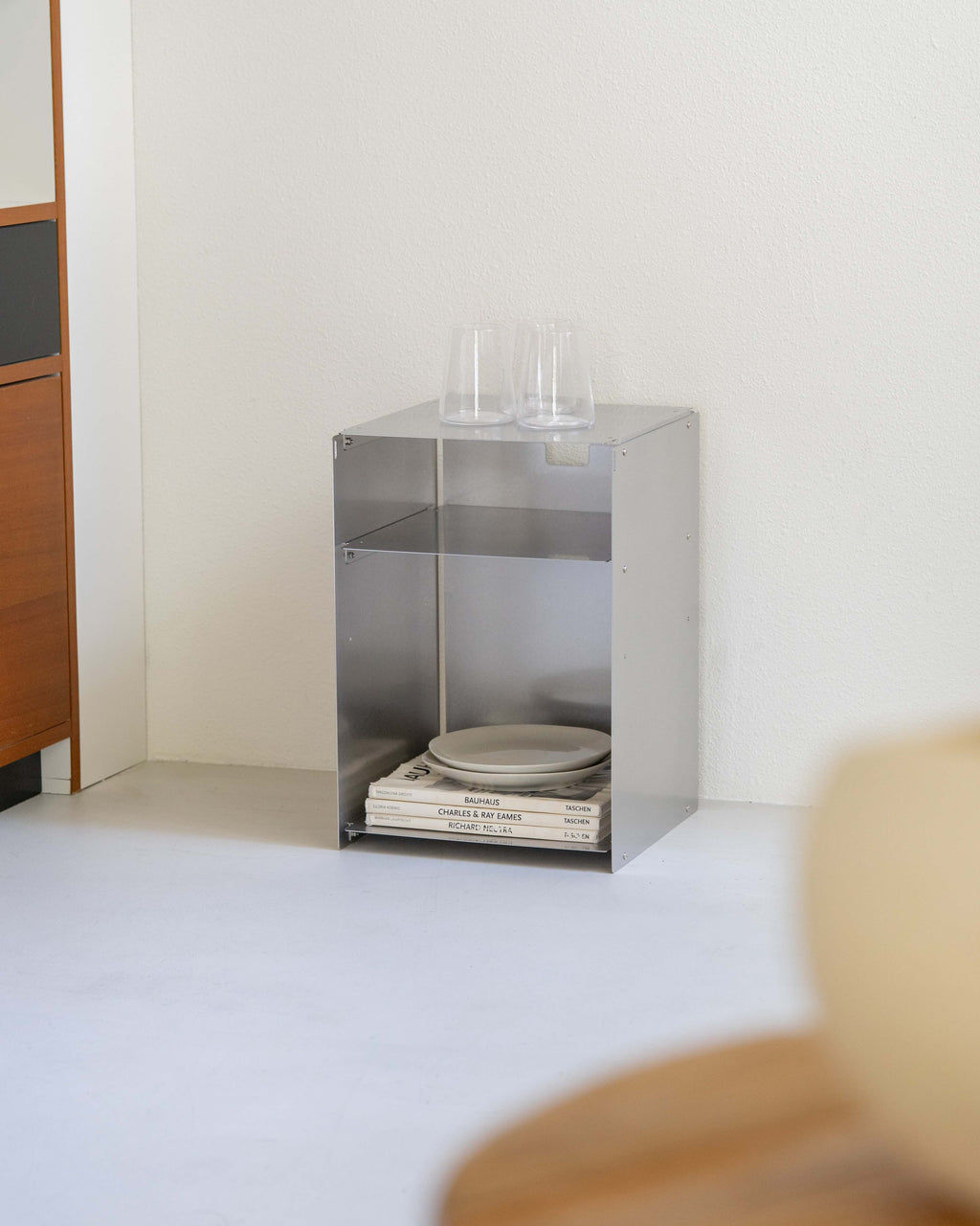 A minimalist stainless steel open shelving cabinet with two tiers, styled with plates and books on the lower shelf and two clear glasses on top, set against a plain off-white wall and floor.