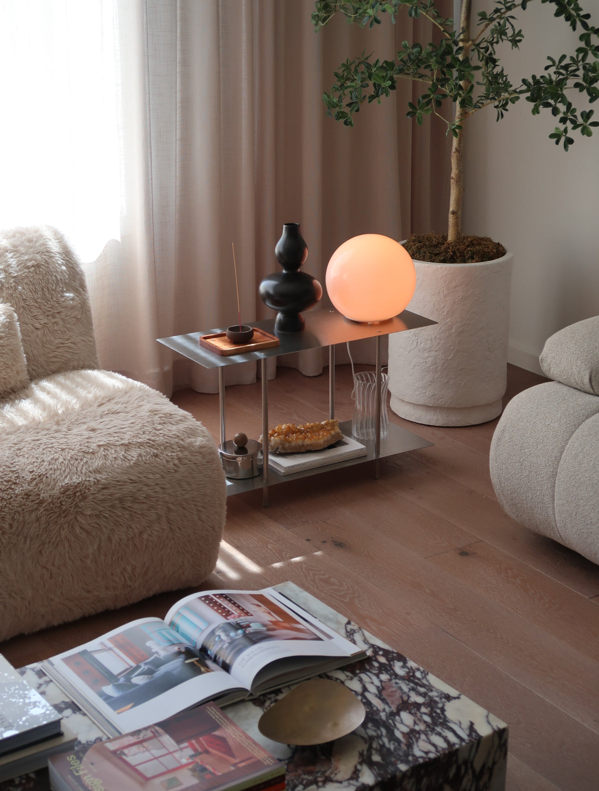 A modern spherical glowing lamp in warm peach-orange sits on a small metal side table in a styled living room. The room features textured beige armchairs, a potted tree, and a marble coffee table in the foreground with open magazines.