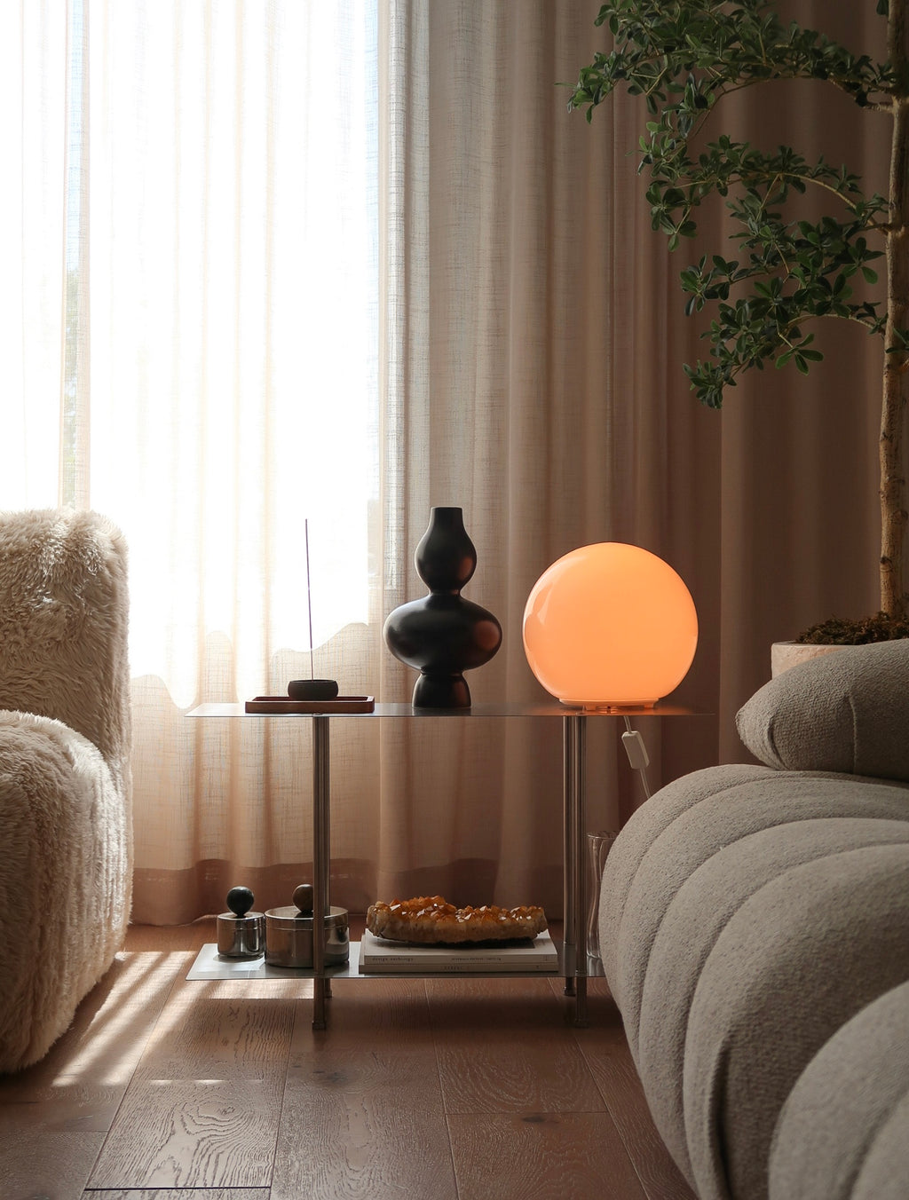 A glowing spherical orange bedside lamp sits on a glass side table in a softly lit living room, next to a textured beige armchair and sofa, with sheer curtains and a potted plant in the background.