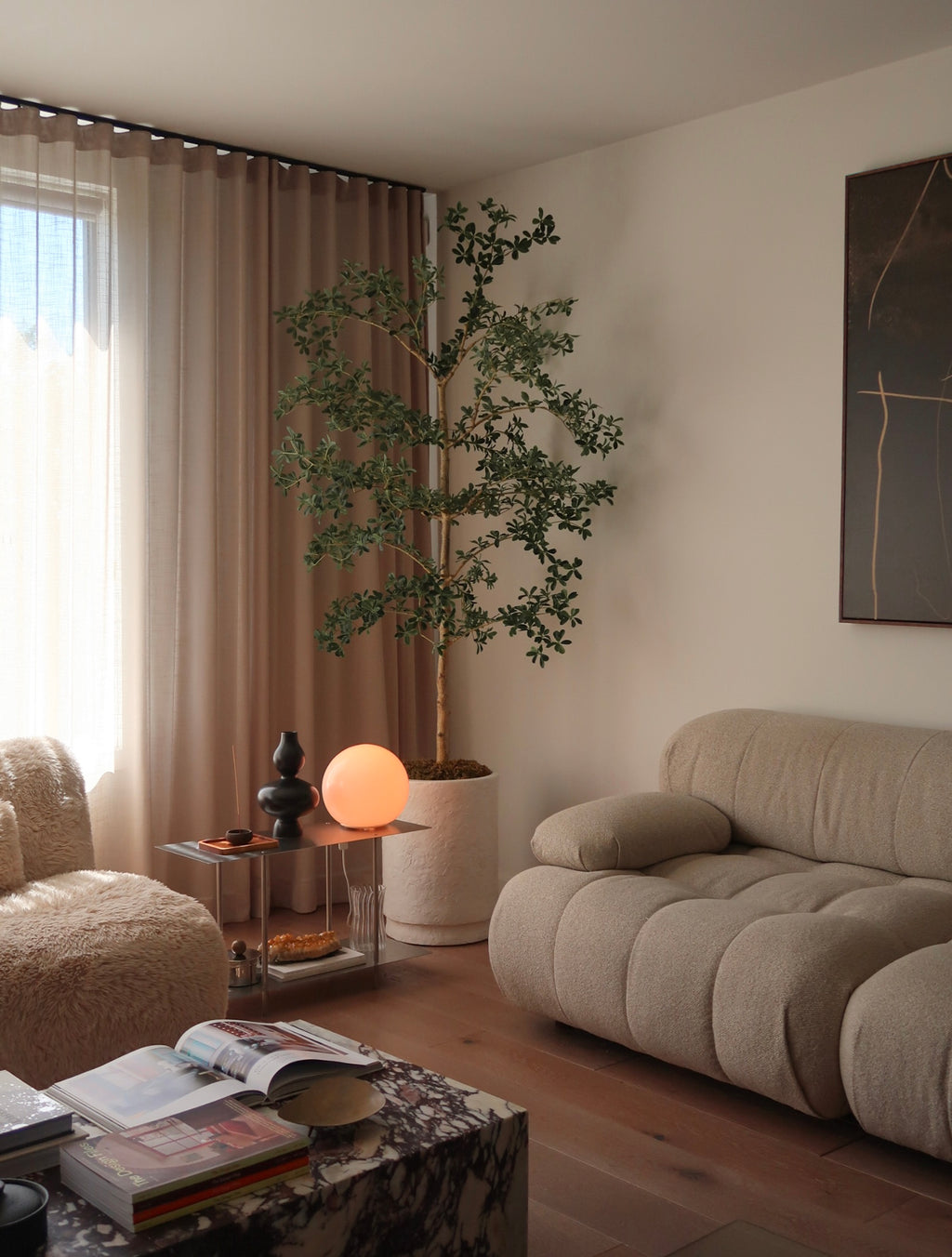 A cozy, modern living room corner featuring a beige tufted sofa, a textured armchair, a potted artificial tree, and a small side table with a glowing orange globe lamp.