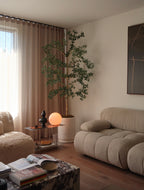A cozy, modern living room corner featuring a beige tufted sofa, a textured armchair, a potted artificial tree, and a small side table with a glowing orange globe lamp.