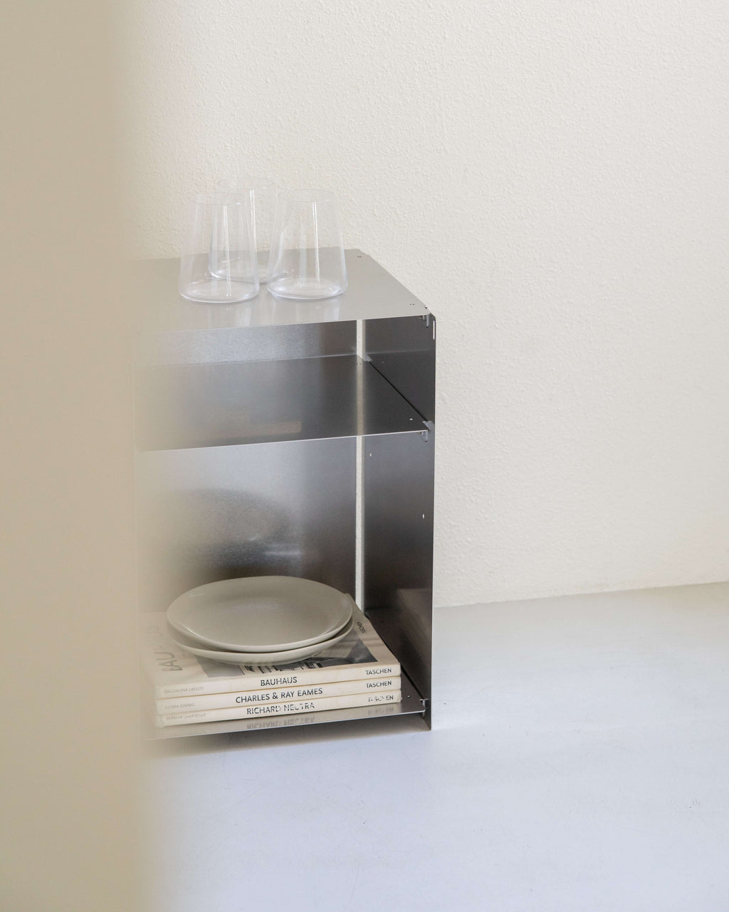 A minimalist stainless steel open shelving unit with two clear glass tumblers on top and stacked plates and books inside, set against a plain off-white wall and floor.