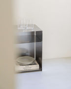 A minimalist stainless steel open shelving unit with two clear glass tumblers on top and stacked plates and books inside, set against a plain off-white wall and floor.