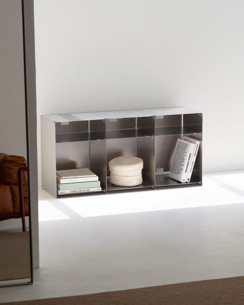A modern dark gray modular storage shelf with multiple cubbies, styled with books, a round ceramic pot, and a magazine, set against a minimalist white wall and floor.