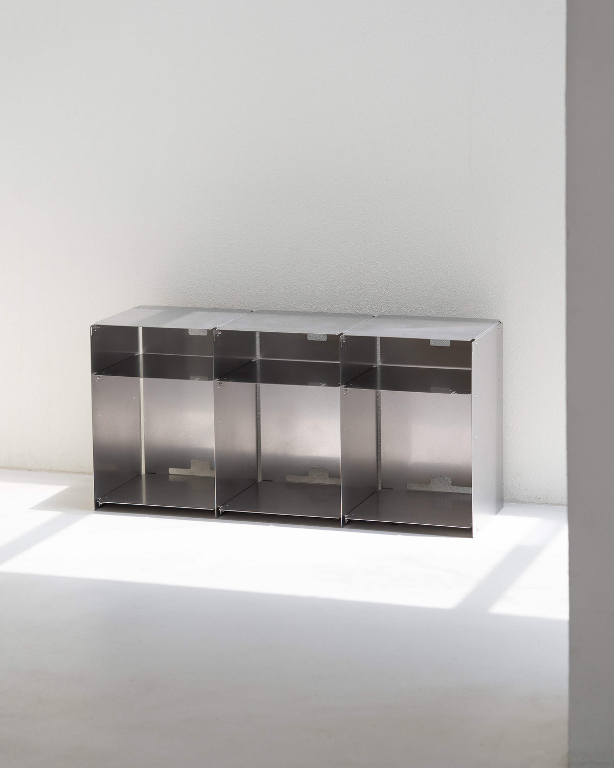 A modern three-compartment metal storage cabinet with a brushed silver finish, set against a minimalist white wall and floor with soft natural light casting shadows.