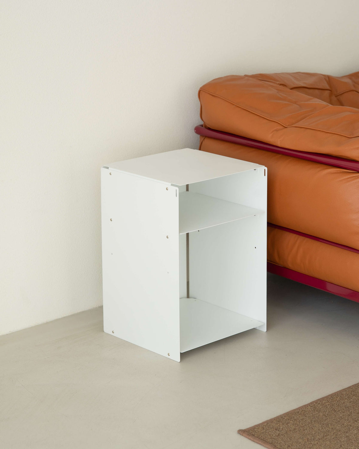 A white wooden two-tier side table or organizer stand placed next to an orange leather sofa in a minimalist room setting.