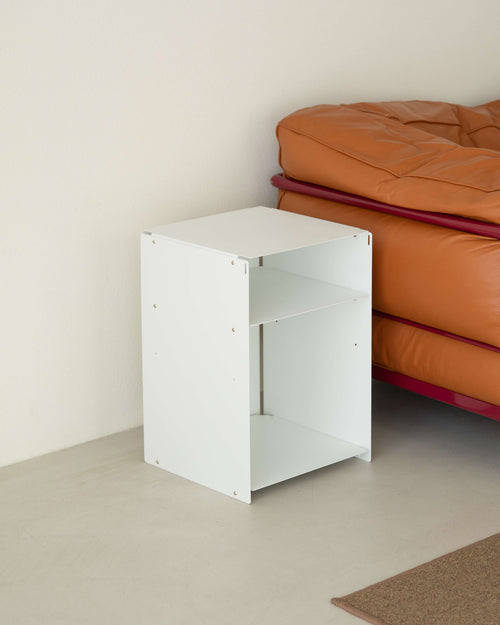 A white wooden two-tier side table or organizer stand placed next to an orange leather sofa in a minimalist room setting.