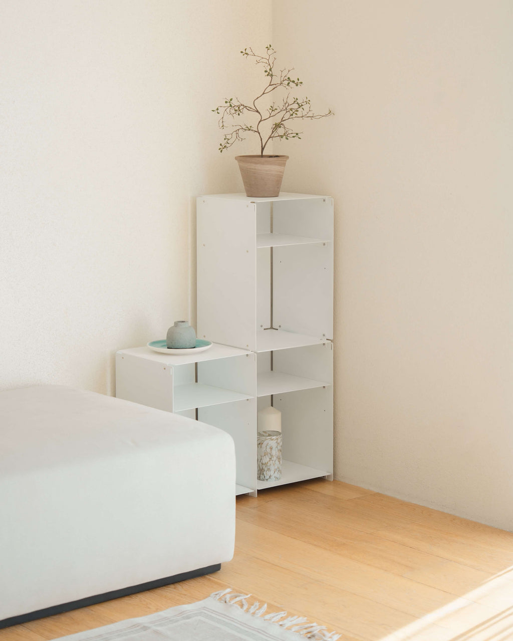 White modular shelving units in a minimalist bedroom corner, with a small side table and a taller open bookcase holding a potted branch, next to a white bed on a light wood floor.