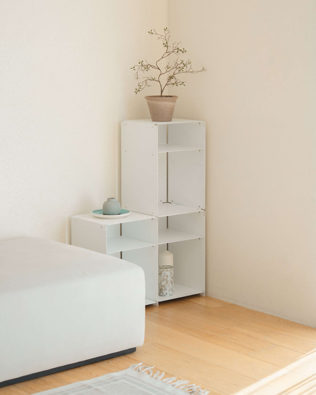 White modular shelving units in a minimalist bedroom corner, with a small side table and a taller open bookcase holding a potted branch, next to a white bed on a light wood floor.