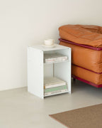 A white wooden two-tier side table or bookcase styled with books and a small white cup, positioned next to an orange leather sofa in a minimalist room.