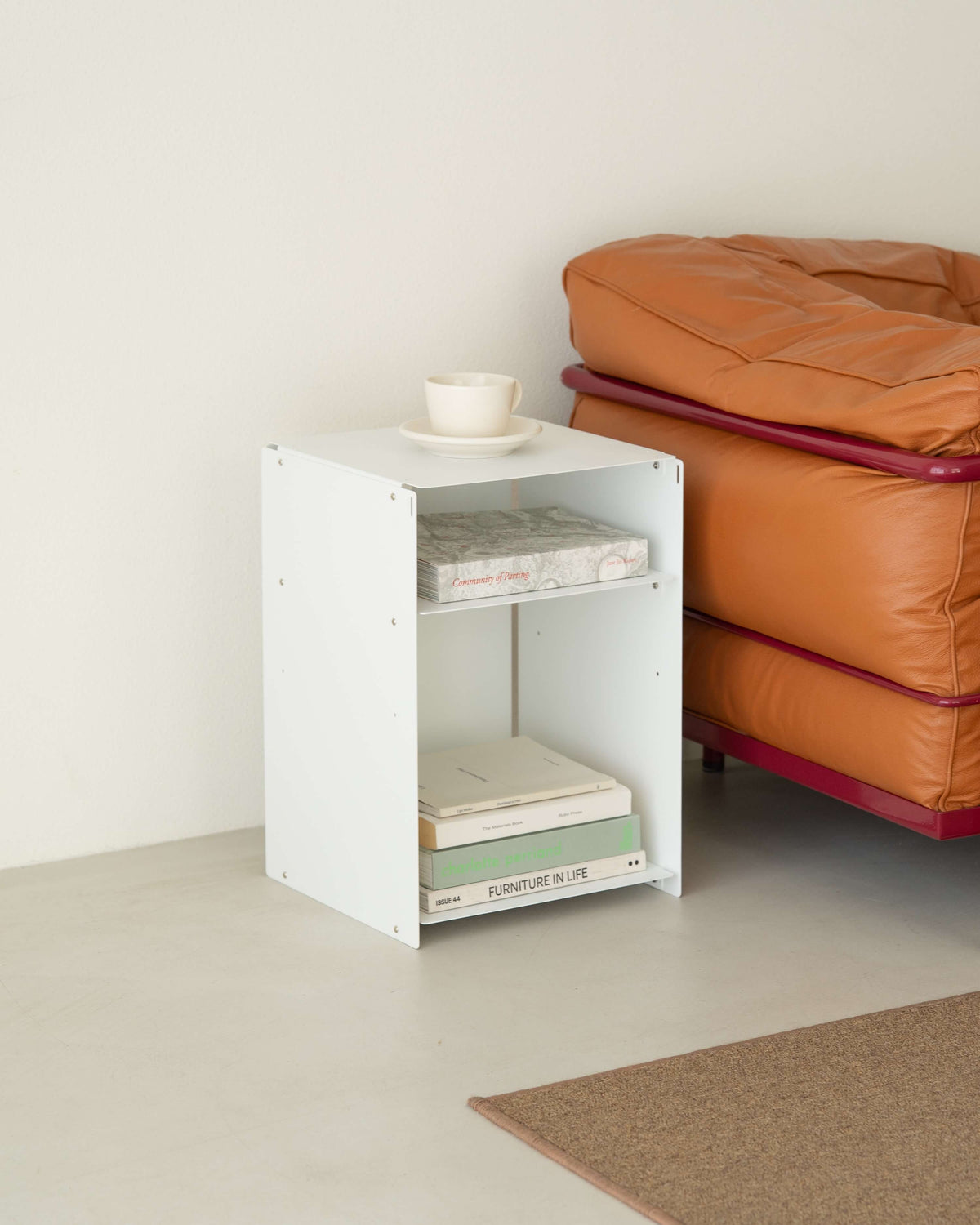 A white wooden two-tier side table or bookcase styled with books and a small white cup, positioned next to an orange leather sofa in a minimalist room.