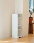 A white modular three-tier bookcase with open shelves, shown in a minimalist room corner with light wood floors, a potted plant on a wooden stool, and a window in the background.