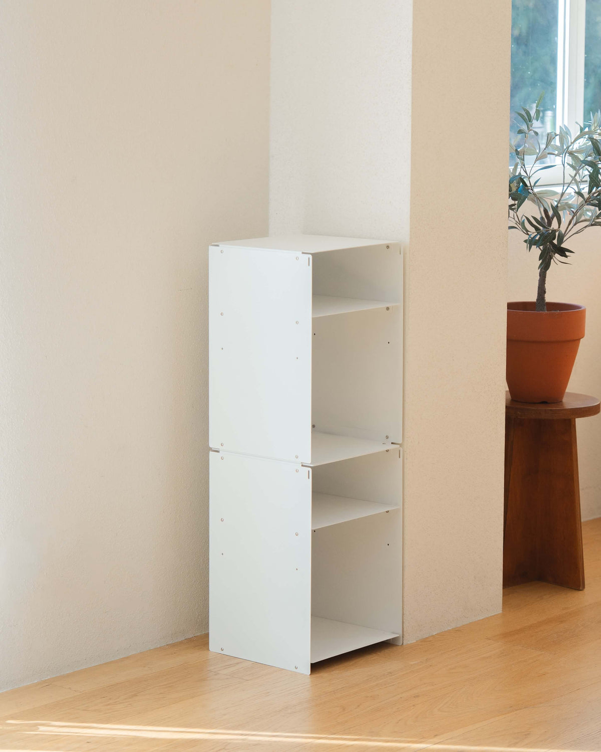 A white modular three-tier bookcase with open shelves, shown in a minimalist room corner with light wood floors, a potted plant on a wooden stool, and a window in the background.